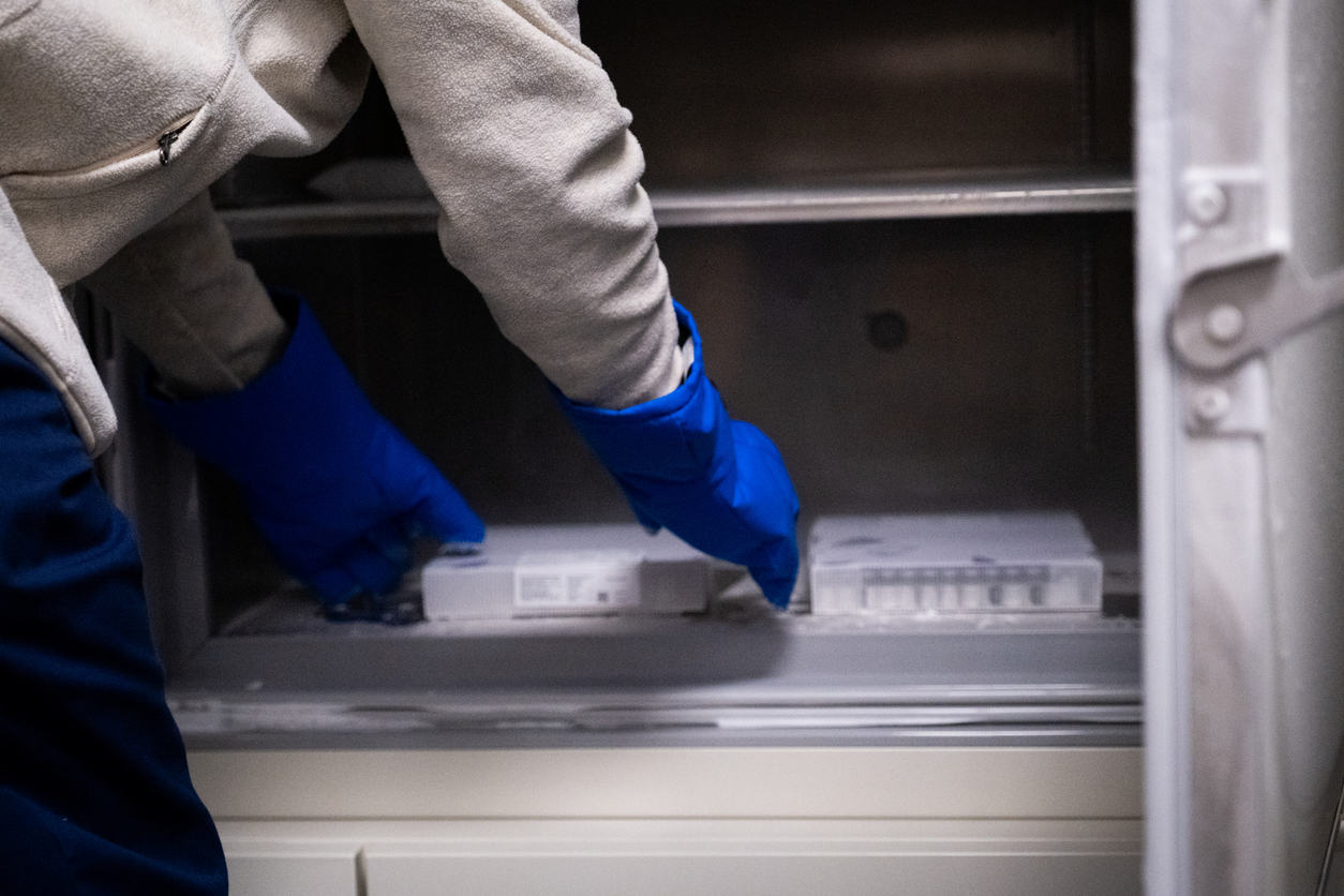 A doctor in a white coat and blue gloves carefully removes a try of the Covid-19 Vaccine out of an industrial refrigerator.