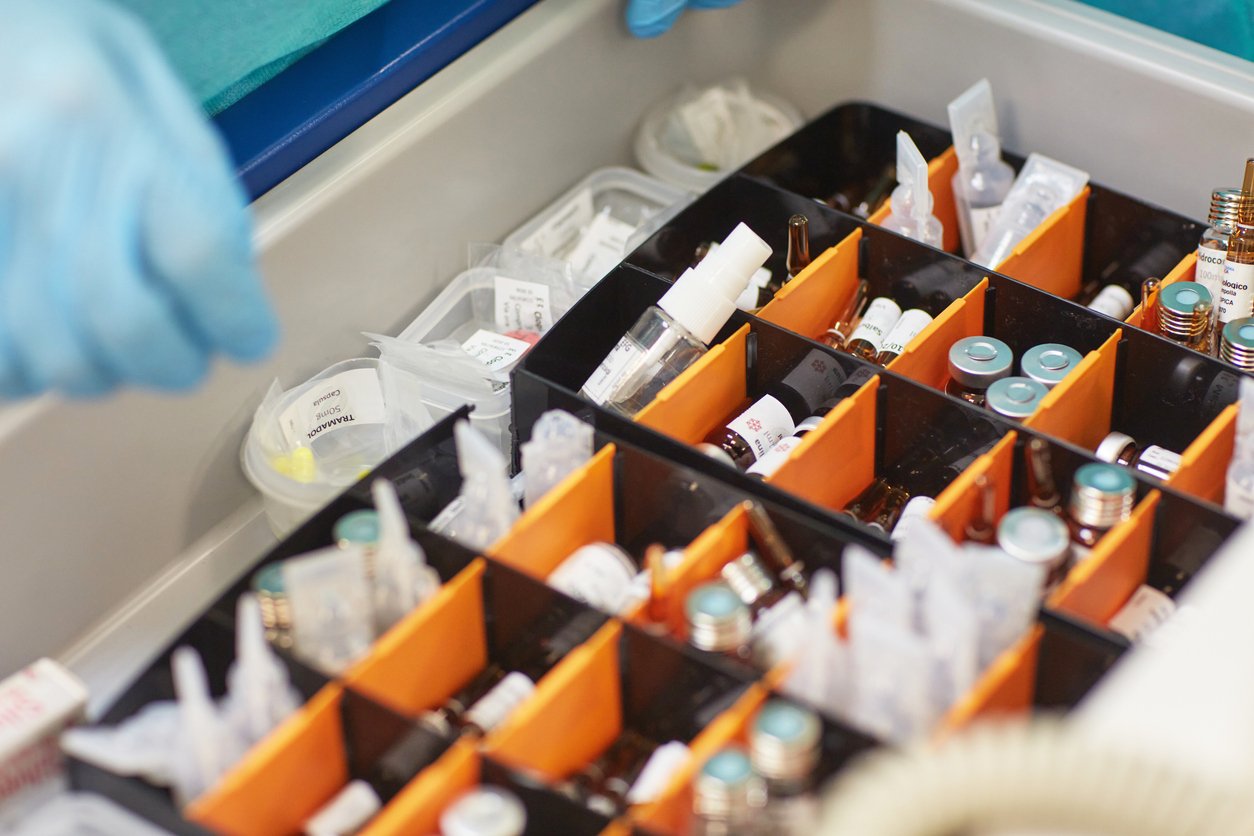Close-up of healthcare worker hands wearing surgical gloves opening crash cart drawer filled with vaccine bottles and ampoules.