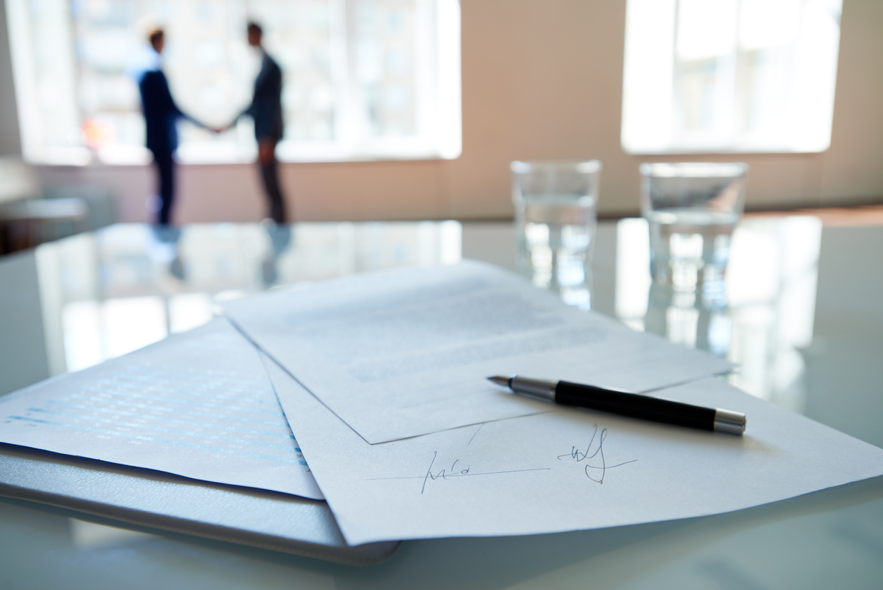 Signed business contract lying on table, business partners shaking hands in the background